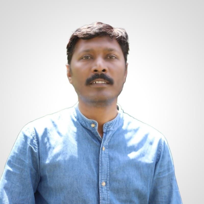 Man with dark hair and mustache wearing a light blue denim shirt against a plain background.