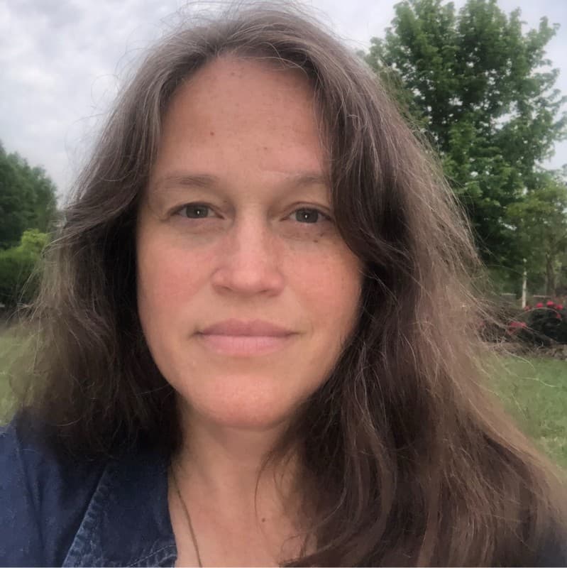 Close-up of a woman with long, wavy brown hair and freckles against a leafy background.